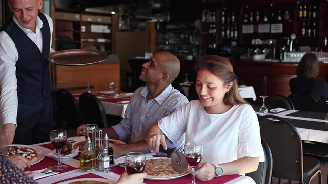 Friendly smiling waiter bringing ordered pizzas to guests of cozy italian restaurant
