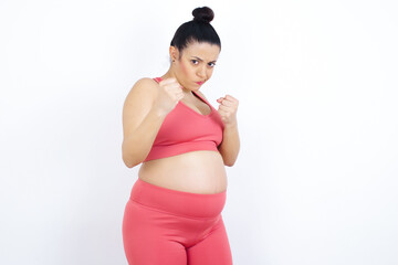 Portrait of attractive young beautiful Arab pregnant woman in sports clothes against white wall holding hands in front of him in boxing position going to fight.