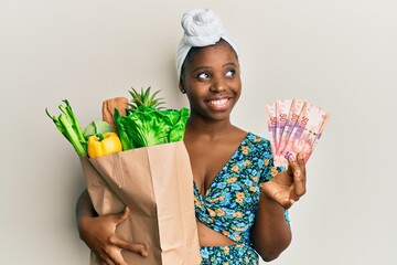 Young african woman holding groceries and 50 rand banknotes smiling looking to the side and staring...