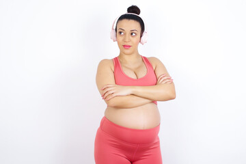 Pleased young beautiful Arab pregnant woman in sports clothes against white wall keeps hands crossed over chest looks happily aside