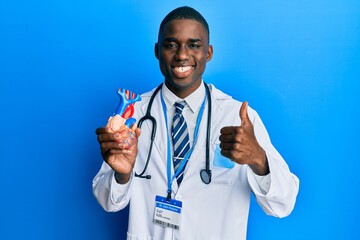 Young african american man wearing doctor uniform holding heart smiling happy and positive, thumb up doing excellent and approval sign