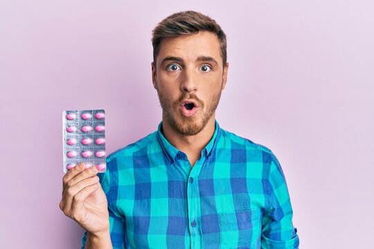 Handsome caucasian man holding pills scared and amazed with open mouth for surprise, disbelief face