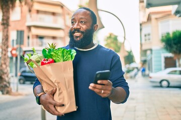 African american man with beard holding paper bag of groceries from supermarket using smartphone