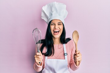 Young hispanic woman wearing baker uniform holding spoon and whisk smiling and laughing hard out...