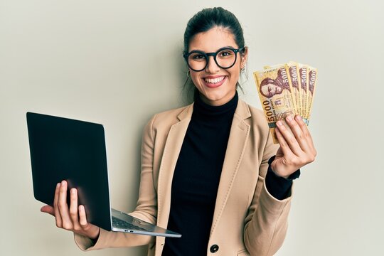 Young hispanic woman wearing business style holding laptop and hungarian forint smiling with a happy and cool smile on face. showing teeth.