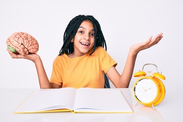 Young african american girl child with braids holding brain while studying for school celebrating achievement with happy smile and winner expression with raised hand