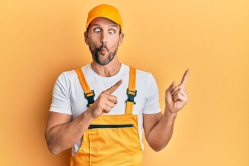 Young handsome man wearing handyman uniform pointing to the side making fish face with mouth and squinting eyes, crazy and comical.