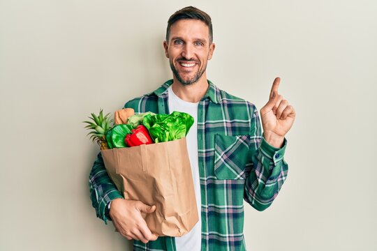Handsome man with beard holding paper bag with groceries cheerful with a smile on face pointing with hand and finger up to the side with happy and natural expression