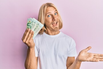 Caucasian young man with long hair holding 20 thai baht banknotes celebrating achievement with happy smile and winner expression with raised hand
