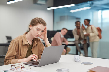 Portrait of stressed young woman using laptop in office or coworking space with group of people in...