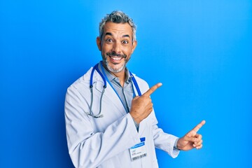 Middle age grey-haired man wearing doctor uniform and stethoscope smiling and looking at the camera pointing with two hands and fingers to the side.