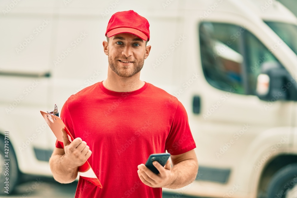 Sticker Young caucasian deliveryman using smartphone and holding clipboard at the city.