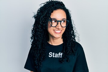 Young hispanic woman with curly hair wearing staff t shirt smiling looking to the side and staring away thinking.