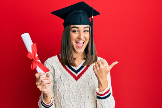 Young brunette girl holding graduate degree diploma pointing thumb up to the side smiling happy with open mouth