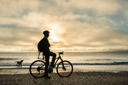 Silhouette image of a cyclist on the bicycle watching the sunrise and dog playing in the water at Milford beach, Auckland