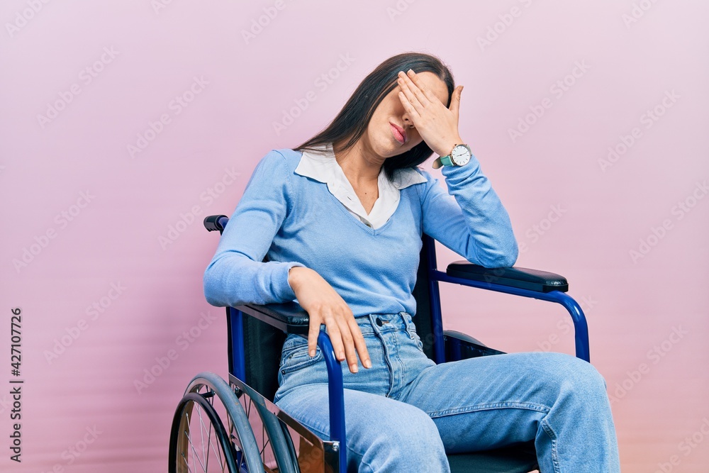 Poster Beautiful woman with blue eyes sitting on wheelchair covering eyes with hand, looking serious and sad. sightless, hiding and rejection concept