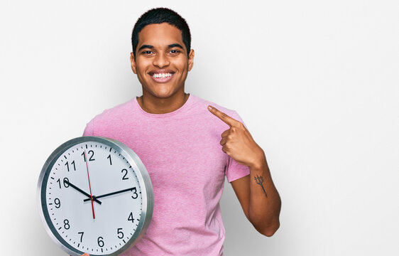 Young handsome hispanic man holding big clock pointing finger to one self smiling happy and proud