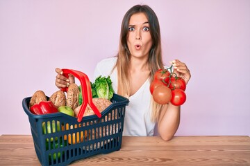 Beautiful caucasian woman holding supermarket shopping basket and tomatoes in shock face, looking skeptical and sarcastic, surprised with open mouth