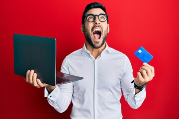 Young hispanic man wearing business style holding laptop and credit card angry and mad screaming...