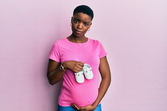 Young african american woman expecting a baby holding shoes skeptic and nervous, frowning upset because of problem. negative person.
