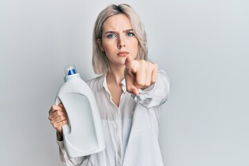 Young blonde girl holding detergent bottle pointing with finger to the camera and to you, confident gesture looking serious