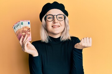 Young blonde girl holding euro banknotes screaming proud, celebrating victory and success very...