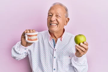 Fotobehang Senior caucasian man holding denture and green apple winking looking at the camera with sexy expression, cheerful and happy face. © Krakenimages.com