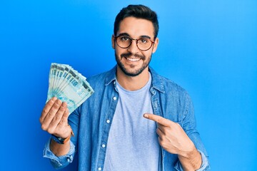 Young hispanic man holding 100 brazilian real banknotes smiling happy pointing with hand and finger