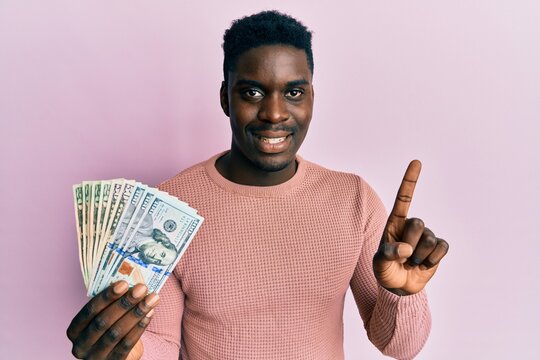 Handsome black man holding dollars smiling with an idea or question pointing finger with happy face, number one