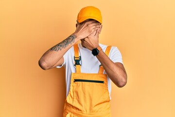 Young handsome african american man wearing handyman uniform over yellow background covering eyes and mouth with hands, surprised and shocked. hiding emotion