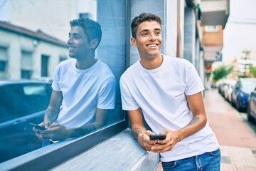 Young latin man smiling happy using smartphone leaning on the wall at the city.