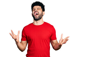 Young arab man with beard wearing casual red t shirt crazy and mad shouting and yelling with aggressive expression and arms raised. frustration concept.