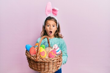Little beautiful girl wearing cute easter bunny ears holding wicker basket with colored eggs afraid and shocked with surprise and amazed expression, fear and excited face.