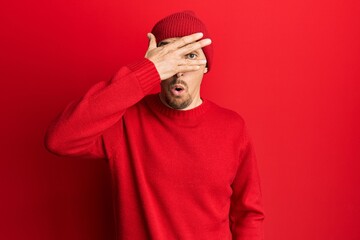 Bald man with beard wearing wool sweater and winter hat peeking in shock covering face and eyes with hand, looking through fingers afraid