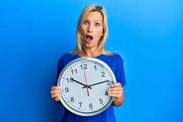 Beautiful middle age blonde woman holding big clock in shock face, looking skeptical and sarcastic, surprised with open mouth