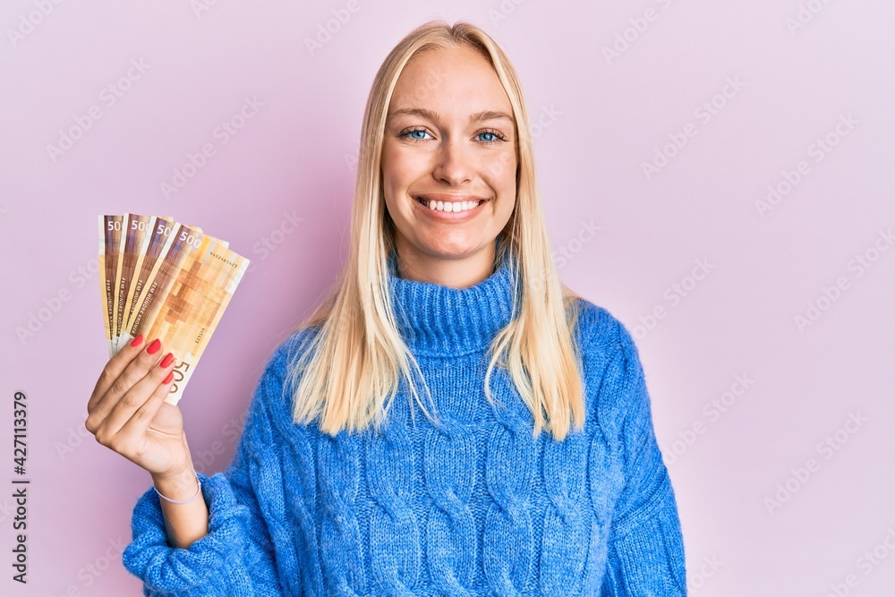 Sticker Young blonde girl holding 100 norwegian krone banknotes looking positive and happy standing and smiling with a confident smile showing teeth