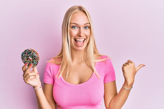 Young blonde girl holding donut pointing thumb up to the side smiling happy with open mouth
