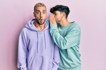 Homosexual gay couple standing together wearing casual clothes hand on mouth telling secret rumor,...