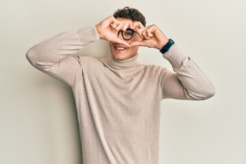 Hispanic young man wearing casual turtleneck sweater doing heart shape with hand and fingers smiling looking through sign