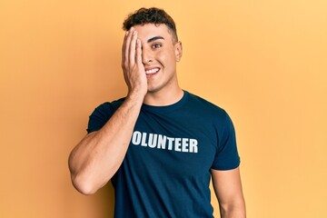 Hispanic young man wearing volunteer t shirt covering one eye with hand, confident smile on face and surprise emotion.