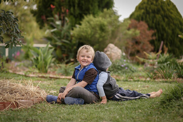 Naklejka na ściany i meble Beautiful blonde brothers sitting back to back on grass in cute garden with happy smiles