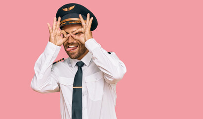 Handsome man with beard wearing airplane pilot uniform doing ok gesture like binoculars sticking tongue out, eyes looking through fingers. crazy expression. - obrazy, fototapety, plakaty