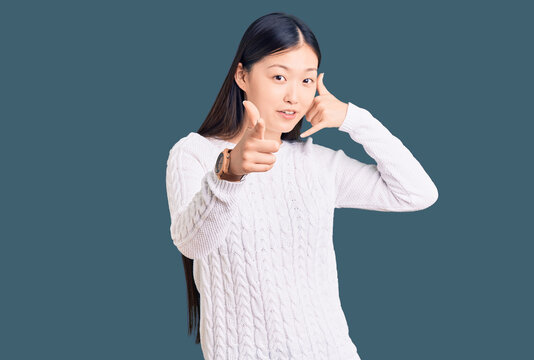 Young beautiful chinese woman wearing casual sweater smiling doing talking on the telephone gesture and pointing to you. call me.