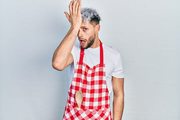 Young hispanic man with modern dyed hair wearing apron surprised with hand on head for mistake,...