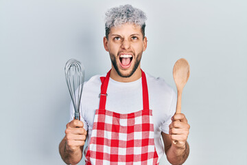 Young hispanic man with modern dyed hair wearing cook apron holding baker whisk and spoon angry and...