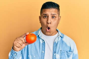 Young latin man holding tangerine scared and amazed with open mouth for surprise, disbelief face