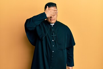 Young latin priest man standing over yellow background covering eyes with hand, looking serious and sad. sightless, hiding and rejection concept