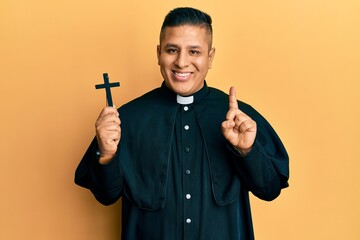 Young latin priest man holding crucifix smiling with an idea or question pointing finger with happy...