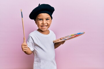 Adorable latin toddler smiling happy wearing artist style using paintbrush and palette over isolated pink background. - obrazy, fototapety, plakaty