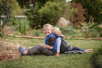 Beautiful blonde brothers sitting back to back on grass in cute garden with happy smiles